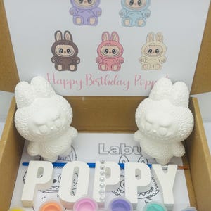 May include: A craft kit in a cardboard box, featuring two white bunny figurines, the name "POPPY" in individual letters, paint pots, and a drawing of bunny characters. The text "Happy Birthday Poppy" is also visible.