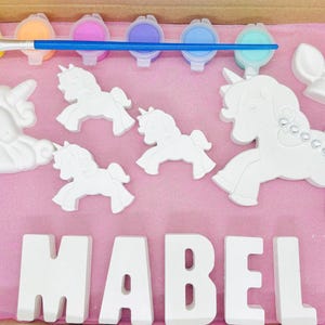 May include: A paint-your-own craft kit with a unicorn theme. The kit includes five paint pots, a paintbrush, and six ceramic pieces: a large unicorn, three smaller unicorns, a unicorn horn, and the name "MABEL" in block letters.