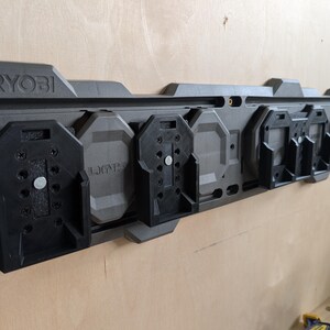 May include: A gray and black RYOBI power tool organizer mounted on a wooden wall. The organizer has multiple black tool holders and the RYOBI logo is visible. The organizer is designed to hold various power tools.