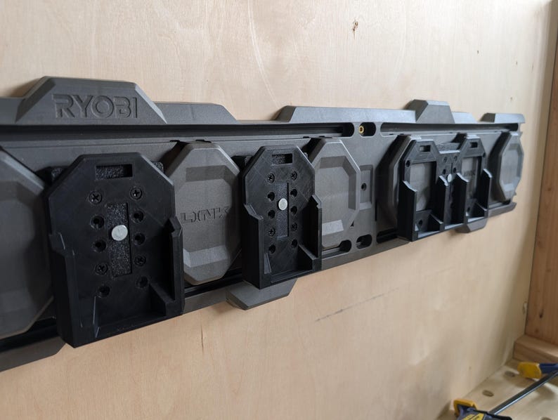 May include: A gray and black RYOBI power tool organizer mounted on a wooden wall. The organizer has multiple black tool holders and the RYOBI logo is visible. The organizer is designed to hold various power tools.