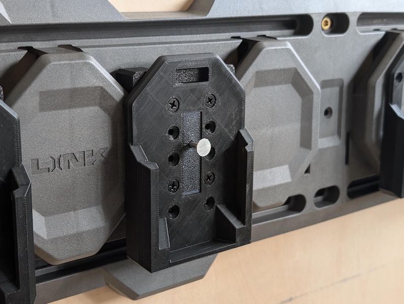 May include: A modular wall-mounted storage system in shades of gray and black. The system features a series of interlocking panels and a black bracket secured with screws. The word "LINK" is embossed on one of the gray panels.