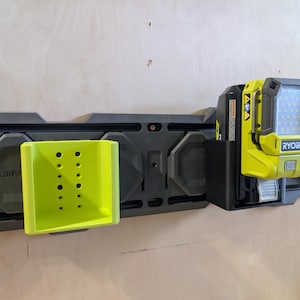 May include: A black and gray wall-mounted storage system with a lime green tool holder and a yellow and black work light. The work light has the brand name "RYOBI" printed on it.