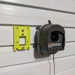 May include: A lime green plastic wall mount bracket with two silver screws and a dark gray battery charger unit attached to a white horizontal slat wall panel. The charger features a black power cord and a warning label with visible text.