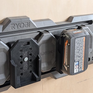 May include: A gray Ryobi power tool organizer with multiple battery holders. A black Ridgid 2Ah lithium battery is attached. The organizer is mounted on a light brown surface.