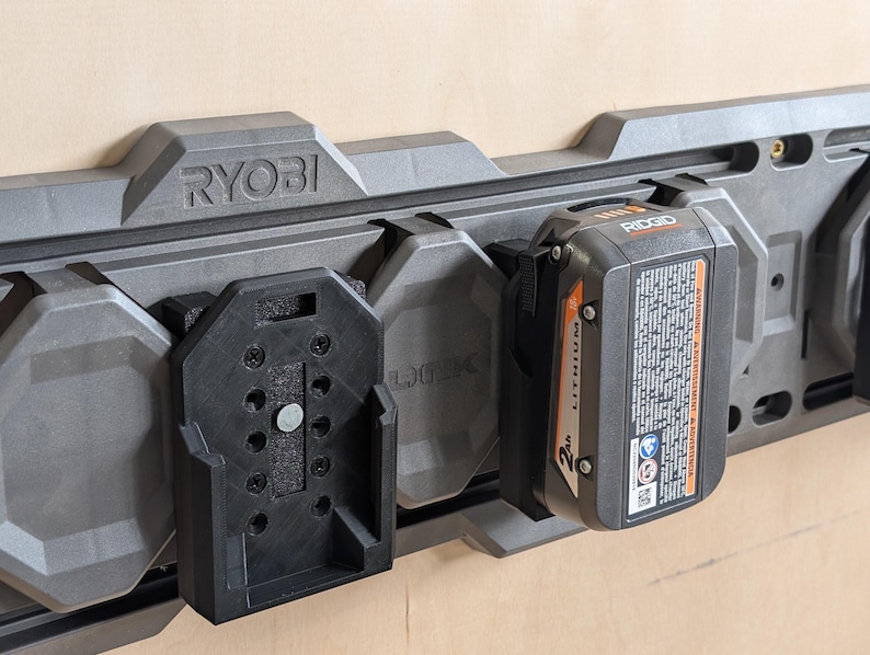 May include: A gray Ryobi power tool organizer with multiple battery holders. A black Ridgid 2Ah lithium battery is attached. The organizer is mounted on a light brown surface.