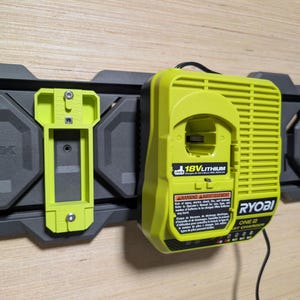 May include: A lime green Ryobi 18V Lithium battery charger mounted on a dark gray wall rail system. The charger features a black power cord, a warning label in English and French, and a secondary lime green mounting bracket attached to the rail.