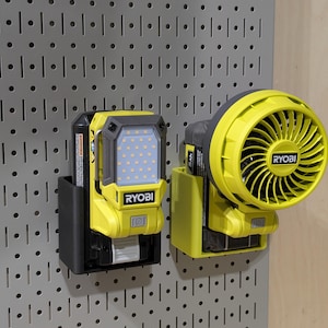 May include: Two yellow and black Ryobi cordless tools mounted on a gray pegboard. One is a work light with a grid of LED lights, and the other is a fan. Both are powered by black battery packs.