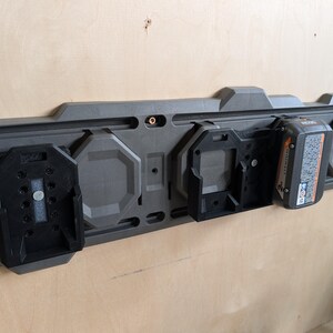 May include: A dark gray wall-mounted power tool battery organizer with black battery holders. One battery is inserted. The organizer is mounted on a light brown wooden wall.