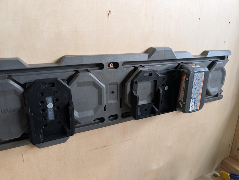 May include: A dark gray wall-mounted power tool battery organizer with black battery holders. One battery is inserted. The organizer is mounted on a light brown wooden wall.