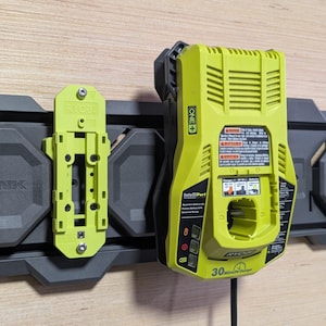 May include: A lime green Ryobi battery charger and mounting bracket attached to a dark gray wall organizer. The charger has a black power cord and a 30-minute charge time indicator.