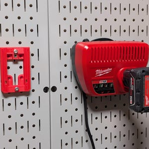 May include: Red Milwaukee M12 battery charger and battery mounted on a pegboard. The charger is red with black accents and the battery is black with red and white labels. A red plastic bracket is also mounted on the pegboard.