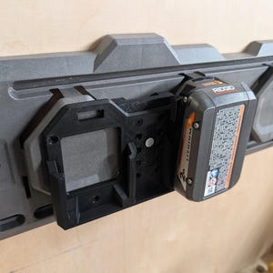 May include: A gray and black power tool battery holder mounted on a light-colored wooden surface. A black battery is inserted into the holder. The battery has the brand name "RIDGID" printed on it.