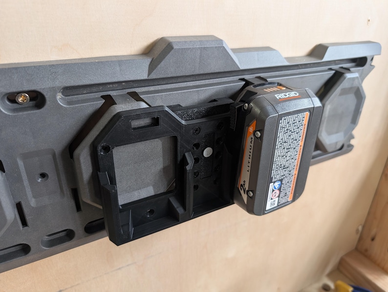 May include: A gray and black power tool battery holder mounted on a light-colored wooden surface. A black battery is inserted into the holder. The battery has the brand name "RIDGID" printed on it.