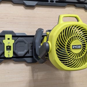 May include: A lime green Ryobi portable fan with a circular grille and top handle, mounted onto a dark gray Ryobi Link wall rail system. The fan features a black center logo and is attached to the rail via a matching lime green locking clip.