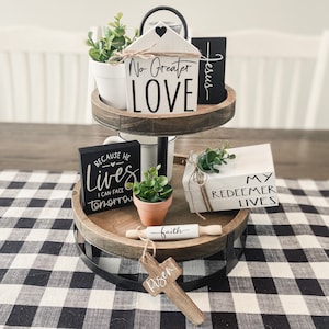 May include: A two-tiered decorative tray with various religious-themed items. The top tier holds a small house-shaped sign with "No Greater LOVE" and a black sign with "Jesus". The bottom tier has a cross, a rolling pin with "faith", and a sign that says "Because He Lives I Can Face Tomorrow".