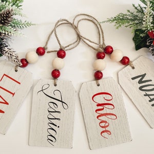 May include: Four wooden gift tags with names in red and black lettering. Each tag is strung with twine and adorned with wooden beads and red spheres. Festive greenery with red berries and pinecones are in the background.