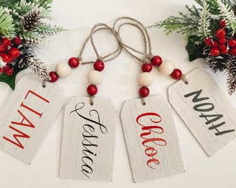 May include: Four wooden gift tags with names in red and black lettering. Each tag is strung with twine and adorned with wooden beads and red spheres. Festive greenery with red berries and pinecones are in the background.