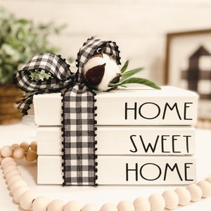 Puede incluir: Tres bloques de madera blancos apilados, con las palabras "HOME SWEET HOME" en letras negras. Una cinta a cuadros en blanco y negro con una mota de algodón y vegetación adorna la parte superior. Una guirnalda de cuentas de madera está en primer plano.