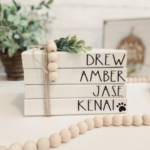 May include: A stack of four white wooden blocks with black lettering spelling out names: DREW, AMBER, JASE, and KENAI. The last block has a paw print. The blocks are tied with twine and adorned with wooden beads and greenery.