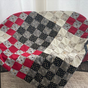 May include: A red, grey, and black checkered quilt with a newspaper print pattern. The quilt is folded and draped over a wicker chair.