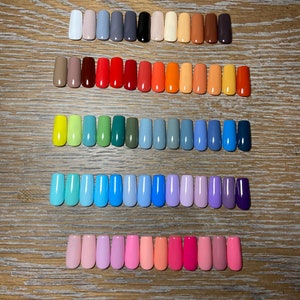 May include: A collection of 50 different colored nail polish colors displayed in rows on a wooden surface. The colors range from light to dark and include shades of red, orange, yellow, green, blue, purple, and pink.