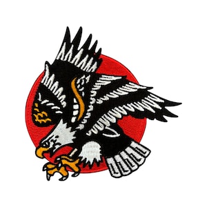Puede incluir: Parche bordado en blanco y negro de un águila con fondo de círculo rojo. El águila está en vuelo con las alas extendidas. La cabeza del águila está girada hacia un lado y su pico está abierto.