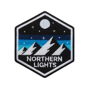 May include: A black and white embroidered patch in a hexagonal shape. The design features a mountain range, a night sky with a full moon, and the text "NORTHERN LIGHTS".