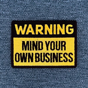 Può includere: Un cartello di avvertimento giallo e nero con il testo "WARNING MIND YOUR OWN BUSINESS" in lettere ricamate nere.