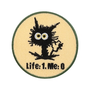 Puede incluir: Parche bordado circular con una criatura negra de dibujos animados con ojos amarillos sobre un fondo amarillo claro. El parche tiene un borde verde y el texto "Life: 1. Me: 0".