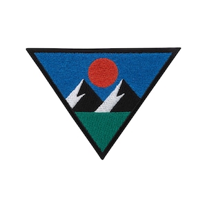 Może przedstawiać: Trójkątna naszywka z motywem górskiego krajobrazu. Ma czarną obwódkę, niebieskie niebo, czerwone słońce, dwie czarne góry z białym śniegiem i zieloną trawę na dole. Odpowiednia do kurtek, toreb lub innych materiałowych przedmiotów.