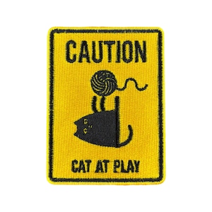 Könnte beinhalten: Ein gelbes Warnschild mit schwarzem Text und einer schwarzen Silhouette einer Katze, die mit einem Wollknäuel spielt. Das Schild lautet "ACHTUNG KATZE IM SPIEL".