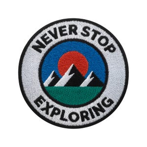 Könnte beinhalten: Runder, gestickter Aufnäher mit dem Text „NEVER STOP EXPLORING“. Das Design umfasst einen blauen Himmel, eine rote Sonne, weiße Berggipfel und grünes Gras. Der Aufnäher hat einen weißen Rand und eine schwarze Umrandung.