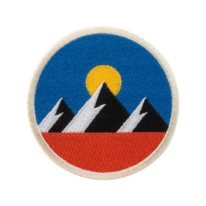 Puede incluir: Parche bordado circular con un diseño de paisaje montañoso. El parche tiene un cielo azul con un sol amarillo, montañas negras con picos blancos y una base roja. El parche tiene un borde de color crema.