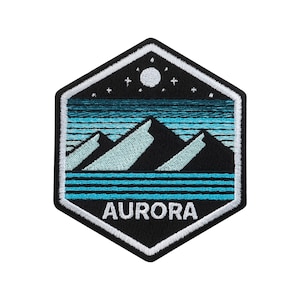 Puede incluir: Parche bordado con forma de hexágono negro. El diseño presenta una cordillera en tonos azules y blancos, con un lago turquesa y un cielo estrellado. La palabra "AURORA" está bordada en blanco.