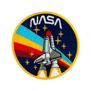Könnte beinhalten: Ein gestickter Aufnäher mit dem NASA-Logo und einem Raumschiff, das vor einem sternenklaren Himmel abhebt, in Blau und Gelb.