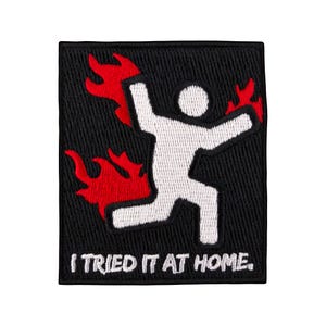 Könnte beinhalten: Ein gestickter Aufnäher in Schwarzweiß mit einer Strichmännchenfigur in Flammen. Der Text "I TRIED IT AT HOME." befindet sich unter der Figur.