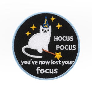 May include: Embroidered patch with a white cat in a wizard hat and wand. The round patch has a black background, blue border, and the text "Hocus Pocus, you've now lost your focus". Includes yellow star accents.
