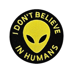Op de afbeelding: Ronde zwarte patch met een gele geborduurde alienkop en de tekst "I DON'T BELIEVE IN HUMANS" in het geel. De alien heeft grote, amandelvormige ogen. Geschikt voor kleding of accessoires.
