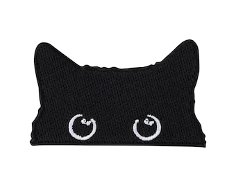 Puede incluir: Un parche bordado negro con forma de parte superior de cabeza de gato con orejas puntiagudas. El parche presenta dos ojos circulares bordados en blanco con pequeños reflejos blancos, situados cerca del borde inferior de la tela negra.