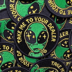May include: A stack of black and green embroidered patches with a cartoon alien smoking a cigarette. The text on the patches reads "Take me to your dealer".