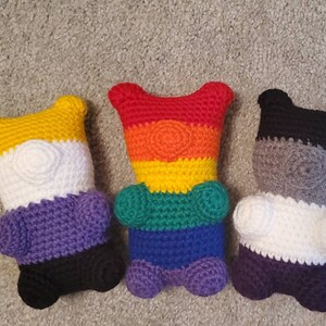 May include: Four crocheted stuffed bears, each with a different colour scheme representing a pride flag. The bears are arranged in a row from left to right: yellow, white, purple, black; red, orange, yellow, green, blue, purple; black, grey, white, purple; and grey, white, black, purple.