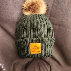May include: A dark green ribbed knit beanie featuring a tan faux fur pom-pom on top. A square tan leather patch is stitched to the front cuff, displaying the letters M and N separated by two crossed hockey sticks in a dark brown print.
