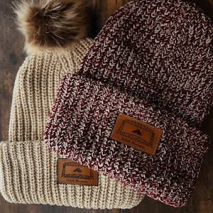 May include: Two knitted beanies with pom poms. One is beige and the other is a mix of burgundy and white. Both have a brown leather patch with the text "Duluth Minnesota" and a graphic of a mountain range.