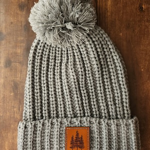 May include: Gray knitted beanie with a large pom pom on top and a brown leather patch with a tree design on the front.