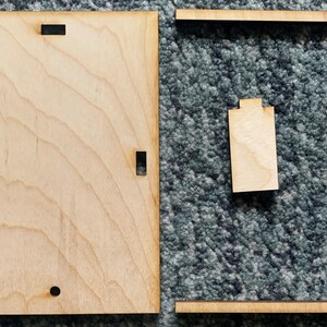 Puede incluir: Dos piezas de madera sin terminar, una es un marco cuadrado y la otra es una pieza rectangular con recortes. Las piezas est&aacute;n sobre una alfombra gris.