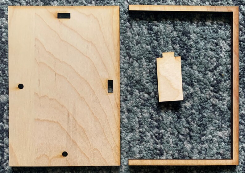 Puede incluir: Dos piezas de madera sin terminar, una es un marco cuadrado y la otra es una pieza rectangular con recortes. Las piezas est&aacute;n sobre una alfombra gris.