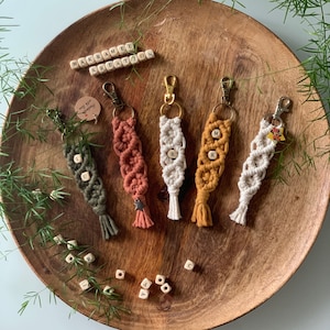 Könnte beinhalten: Fünf bunte Makramee-Schlüsselanhänger mit Holzbuchstabenperlen. Die Schlüsselanhänger hängen an einem Holztablett mit den Worten "Makrame Kreation" in Holzbuchstaben.