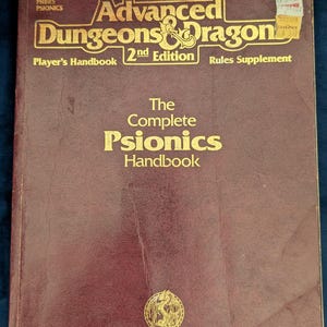 May include: A maroon Advanced Dungeons & Dragons 2nd Edition Player's Handbook Rules Supplement titled The Complete Psionics Handbook. The cover features gold text, a dragon logo, and a small circular TSR emblem at the bottom center.