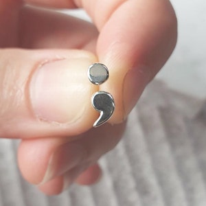 May include: A pair of silver semicolon earrings. The earrings are small, with a polished, reflective surface. The design is a minimalist representation of the punctuation mark.