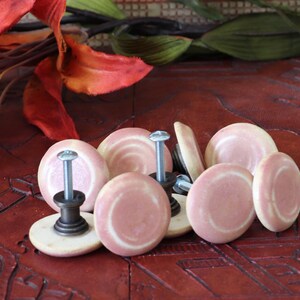May include: A collection of pink and cream-colored cabinet knobs with silver screws. The knobs have a circular design with a lighter pink center and a cream-colored edge. They are arranged on a dark red, carved wooden surface.